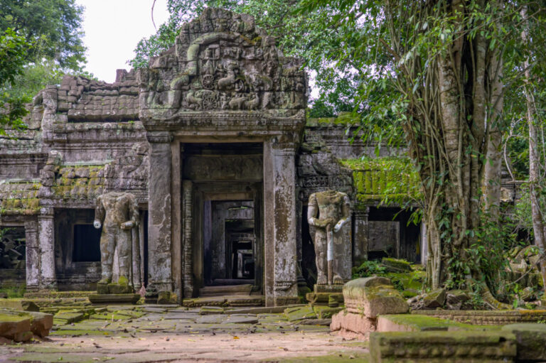 South entrance of Preah Khan temple in Angkor, featuring two headless statues on either side of the ancient stone gateway, surrounded by lush jungle.